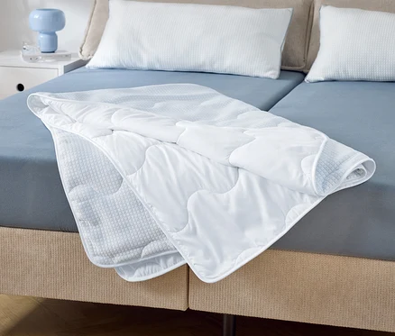 Auf einem Doppelbett mit blauen Laken liegt eine teilweise gefaltete, gesteppte, weiße Decke. Zwei weiße Kissen liegen am Kopfende.
