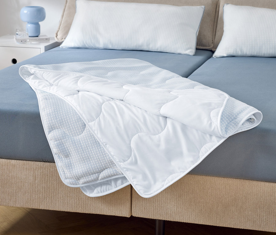 Auf einem Doppelbett mit blauen Laken liegt eine teilweise gefaltete, gesteppte, weiße Decke. Zwei weiße Kissen liegen am Kopfende.