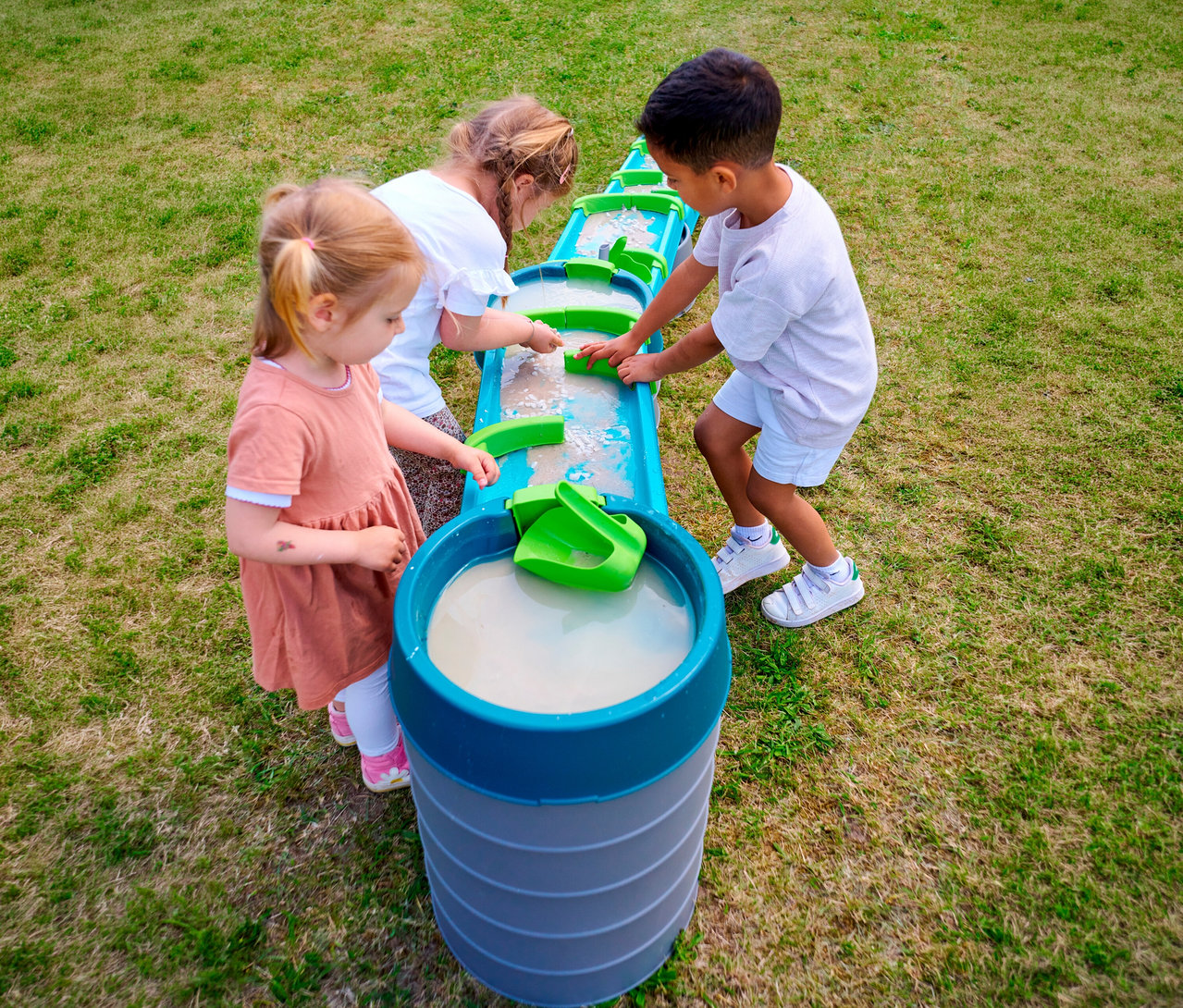 Drei Kinder spielen mit der AquaPlay »Mudway STREAM« Matschbahn im Gras und bewegen grüne Elemente im Wasser.