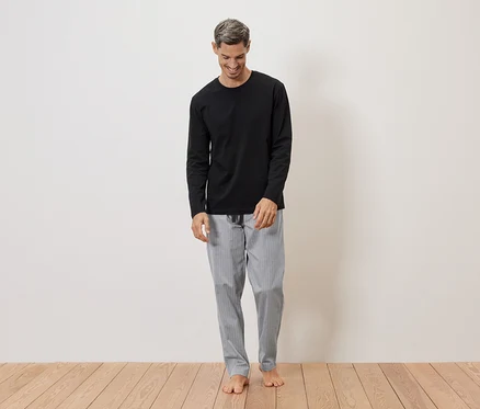 Mann mit grauen Haaren trägt ein schwarzes Langarmshirt und eine grau gestreifte Pyjamahose, barfuß vor einer weißen Wand.