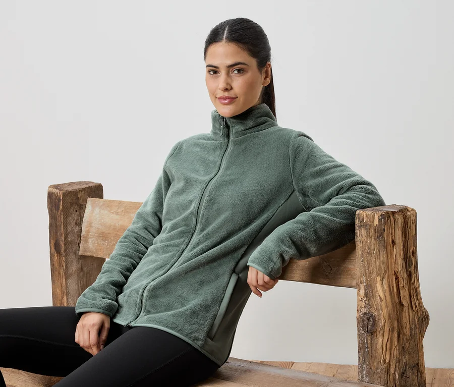 Frau sitzt auf einer Holzbank und trägt eine grüne Fleecejacke und schwarze Leggings.