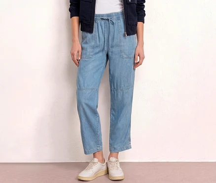 Frau trägt hellblaue Jeans, weiße Schuhe und eine dunkelblaue Jacke vor einer weißen Wand.