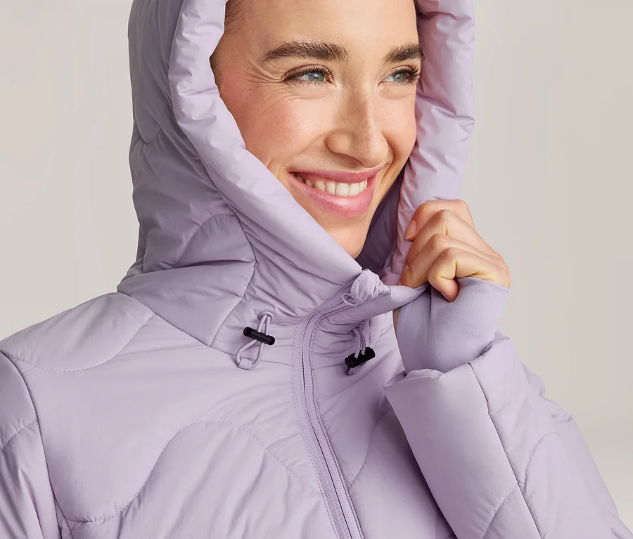 Lachende Frau in einer hellvioletten Steppjacke mit Kapuze, die den Kragen festhält.