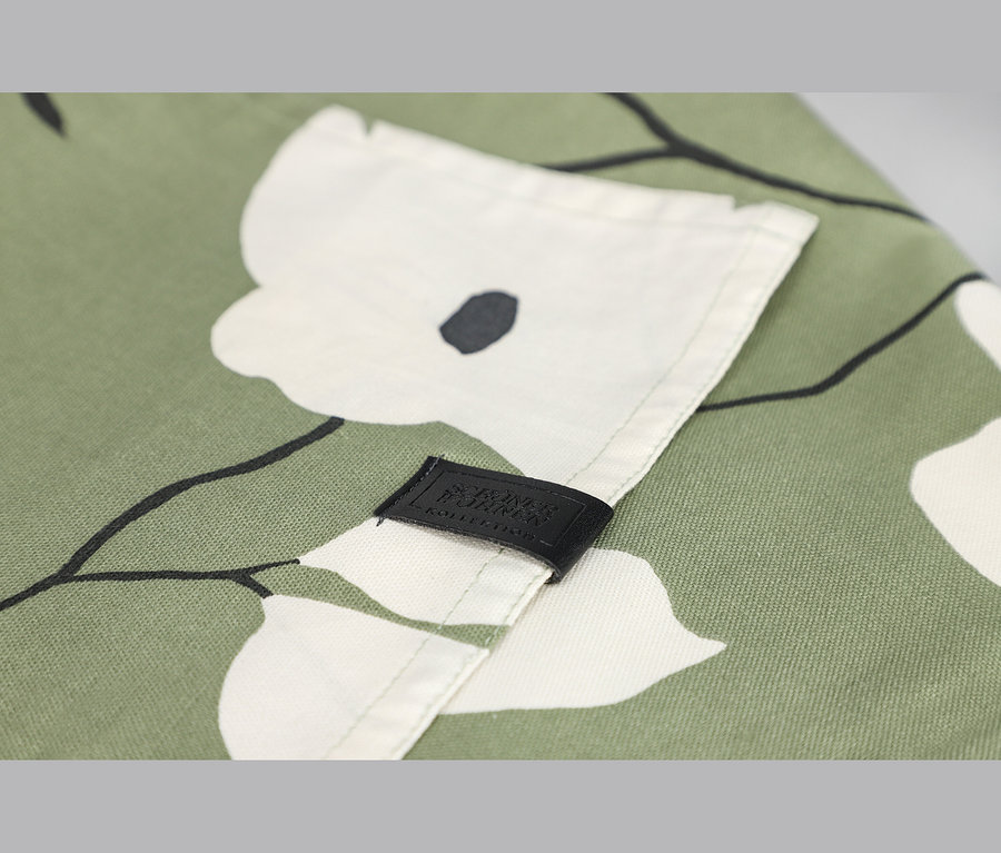 Nahaufnahme eines grünen Stoffes mit weißem Blumenmuster und schwarzen Details. Ein schwarzes Etikett mit der Aufschrift „SCHÖNER WOHNEN KOLLEKTION“ ist an einer gefalteten Stoffkante angenäht.