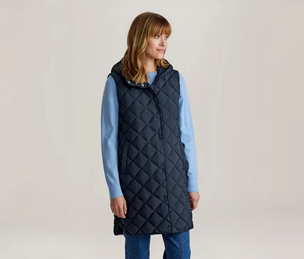 Frau mit schulterlangem braunem Haar und Pony, trägt eine lange, gesteppte, marineblaue Kapuzenweste über einem hellblauen Langarmshirt und blauen Jeans, blickt nach rechts.