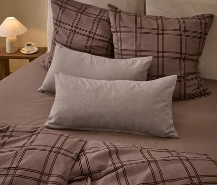Ein Bett mit brauner karierter Bettdecke und zwei passenden großen Kissen, zwei einfarbigen hellgrauen rechteckigen Kissen und einem braunen Spannbettlaken. Eine weiße Lampe und eine Tasse Kaffee stehen auf einem Holznachttisch.