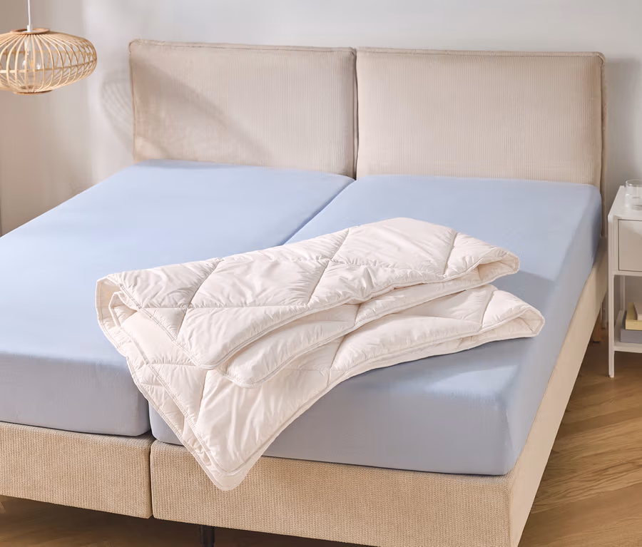 Ein Doppelbett mit beigem Kopfteil und zwei hellblauen Spannbettlaken. Eine gefaltete, cremefarbene Steppdecke liegt auf der rechten Bettseite.