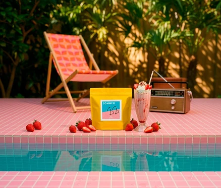 Ein gelber Heilandt Sommer Omni-Roast Kaffeebeutel, ein rosa Erdbeer-Milchshake mit Sahne und frische Erdbeeren stehen auf rosafarbenen Poolfliesen. Im Hintergrund sind ein gemusterter Liegestuhl, ein Vintage-Radio und grüne Pflanzen zu sehen.