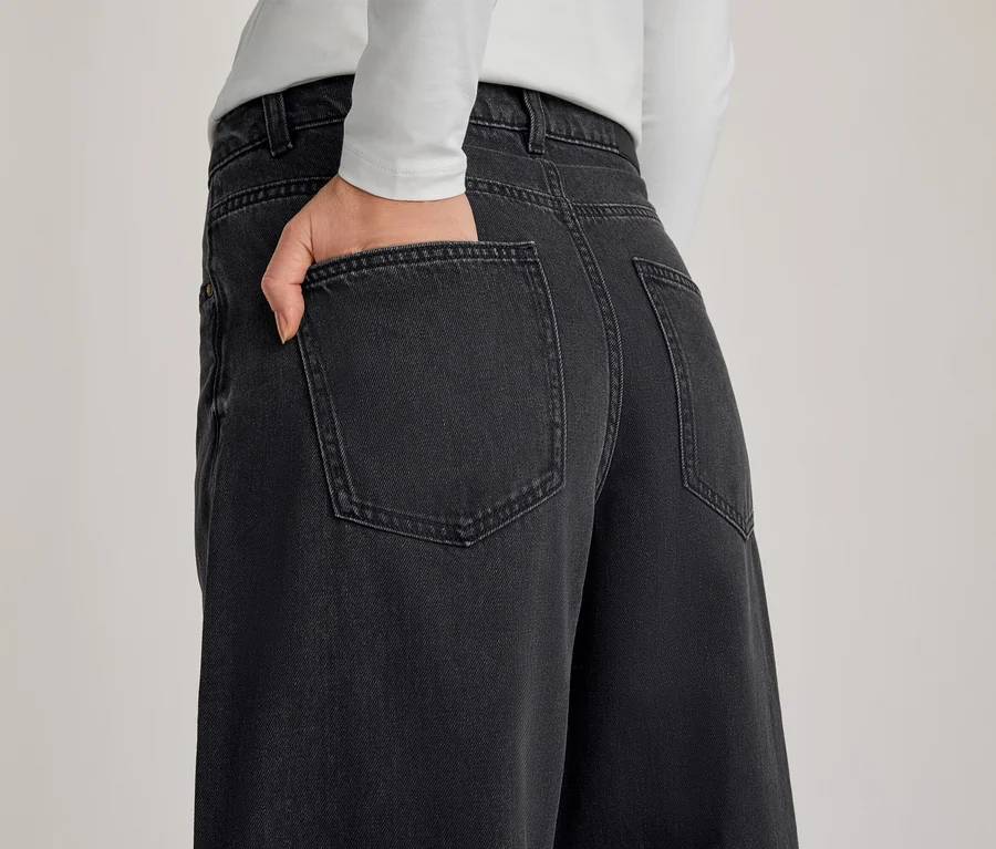 Person von hinten, trägt eine dunkelgraue Wide-Leg-Jeans und ein weißes Langarmshirt, eine Hand steckt in der Gesäßtasche der Jeans.