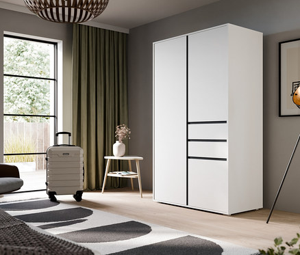 Ein modernes Schlafzimmer mit einem weißen Kleiderschrank, einem Bett mit grauer Decke und einem schwarz-weißen Teppich. Links befinden sich ein großes Fenster mit grünen Vorhängen, ein brauner Sessel, ein hellgrauer Koffer und ein kleiner Beistelltisch mit Blumen. Rechts stehen eine schwarze Stehlampe und gerahmte abstrakte Kunst.