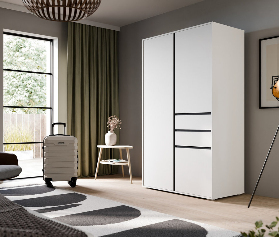 Ein modernes Schlafzimmer mit einem weißen Kleiderschrank, einem Bett mit grauer Decke und einem schwarz-weißen Teppich. Links befinden sich ein großes Fenster mit grünen Vorhängen, ein brauner Sessel, ein hellgrauer Koffer und ein kleiner Beistelltisch mit Blumen. Rechts stehen eine schwarze Stehlampe und gerahmte abstrakte Kunst.