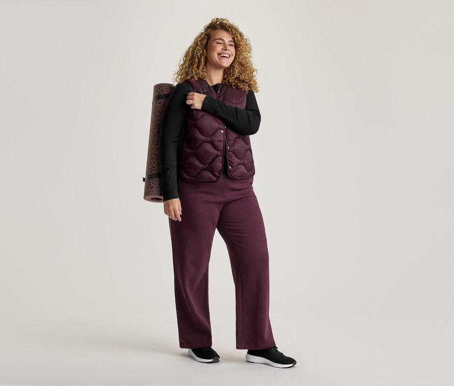 Lachende Frau mit Locken, trägt ein schwarzes Langarmshirt, eine gesteppte bordeauxrote Weste und weite Hosen, eine Yogamatte mit Leopardenmuster über der Schulter.