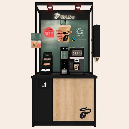 Eine schwarz-holzfarbene Selbstbedienungs-Kaffeestation mit Tchibo Logo. Ein großes Bild zeigt eine Hand, die einen Kaffeebecher hält, mit dem Text „GROSSER KAFFEE, KLEINER PREIS“ und „Fairer Genuss TO GO!“. Eine schwarze Kaffeemaschine mit Touchscreen-Display gibt Kaffee in einen Markenbecher aus. Links daneben befinden sich ein Spender für Rührstäbchen, Zucker und Deckel sowie eine Menütafel. Der untere Teil der Station hat eine Müllöffnung und ein großes Kaffeebohnen-Logo.