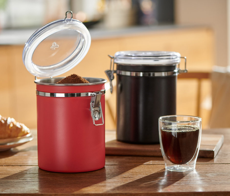 Auf einem Holztisch stehen eine offene rote und eine geschlossene schwarze Aromadose neben einem Glas Kaffee und einem Teller mit einem Croissant.
