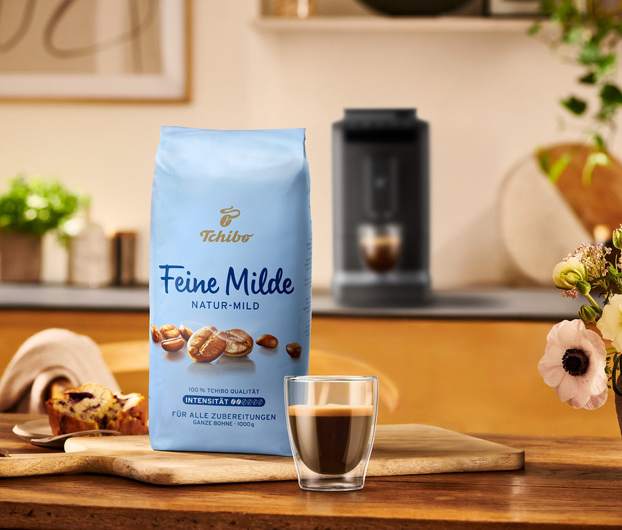 Eine Packung Tchibo Feine Milde Kaffee steht neben einer Tasse Espresso und einem Stück Kuchen auf einem Holztisch.