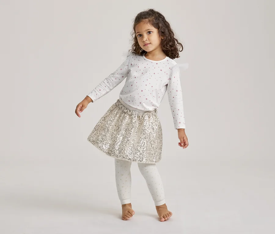Ein kleines Mädchen mit lockigem Haar steht barfuß und trägt ein weißes Langarmshirt mit rosa Sternen, einen goldenen Paillettenrock und gepunktete Leggings.