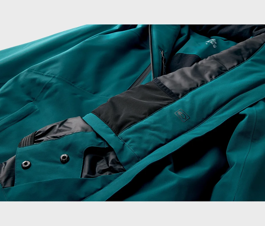 Nahaufnahme einer tealfarbenen Funktionsjacke mit schwarzem Futter, sichtbarem Reißverschluss, zwei Druckknöpfen und einem geprägten Brillenlogo am Kragen.