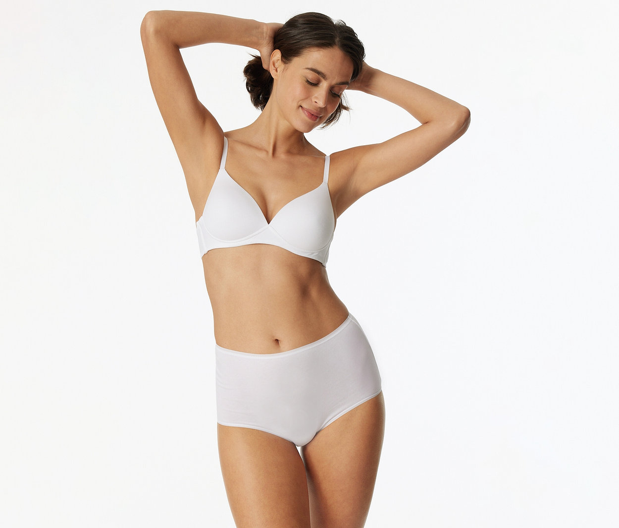 Frau mit dunklem Haar, die einen weißen BH und einen weißen High-Waist-Slip trägt, die Arme hinter dem Kopf verschränkt und lächelnd die Augen geschlossen hat.