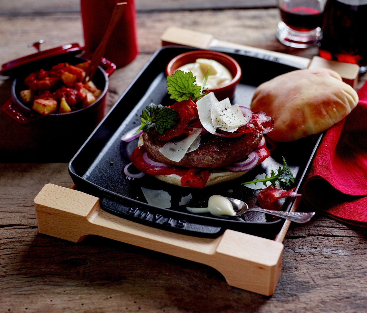 Ein Burger mit roter Sauce, Zwiebeln und Käse auf einem STAUB Teller mit Holzunterlage, daneben Kartoffeln in einem Topf und Rotwein.