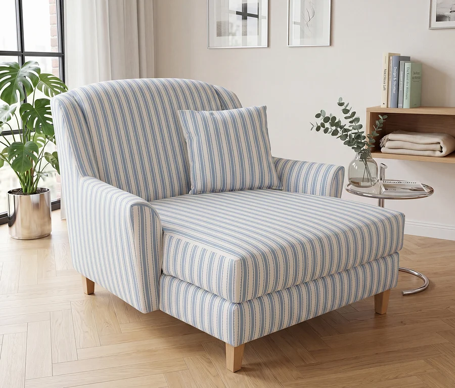 Ein hellblau-weiß gestreifter Chaiselongue mit passendem Kissen steht auf einem hellen Parkettboden in einem Wohnzimmer. Links sind ein großes Fenster mit weißen Vorhängen und eine Topfpflanze zu sehen, rechts ein Holzregal mit Büchern und einer Decke sowie ein Beistelltisch aus Glas mit einer Eukalyptusvase.