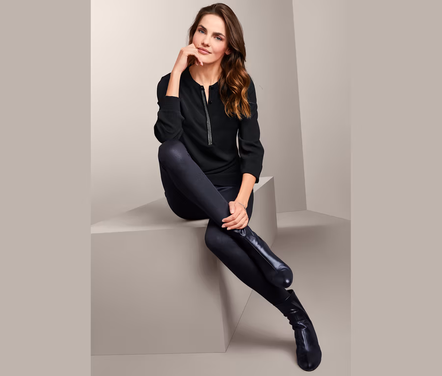 Frau sitzt in einer verzierten Tunikabluse im Materialmix, schwarzen Leggings und Stiefeln.