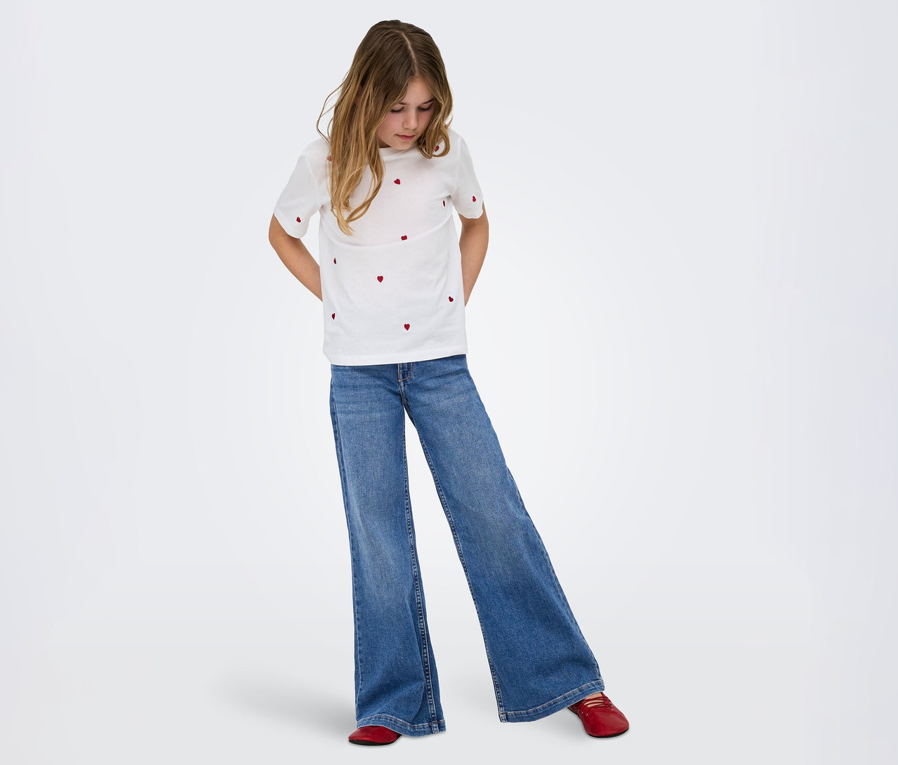 Ein junges Mädchen mit langen braunen Haaren trägt ein weißes T-Shirt mit roten Herzmustern, weite blaue Jeans und rote flache Schuhe und blickt nach unten.