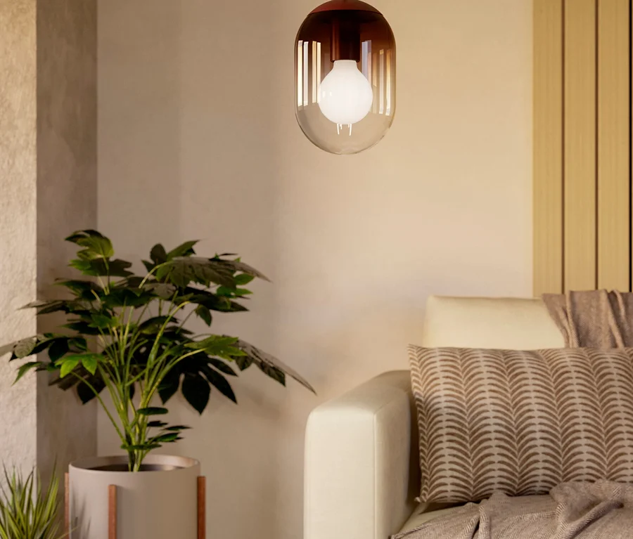 Eine moderne Pendelleuchte mit klarem und rötlich-braunem Glasschirm hängt über einem runden Holztisch in einer gemütlichen Wohnzimmerecke. Rechts steht ein helles Sofa mit gemustertem Kissen und einer Decke. Auf dem Tisch liegen eine offene Zeitschrift, eine dunkle Schale und eine gemusterte Kerze. Links stehen zwei Topfpflanzen.