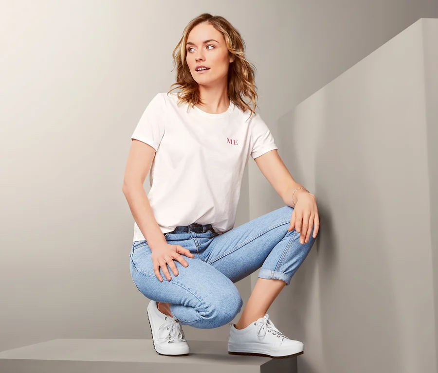 Eine Frau in einem weißen T-Shirt und blauen Jeans hockt auf einem grauen Würfel.