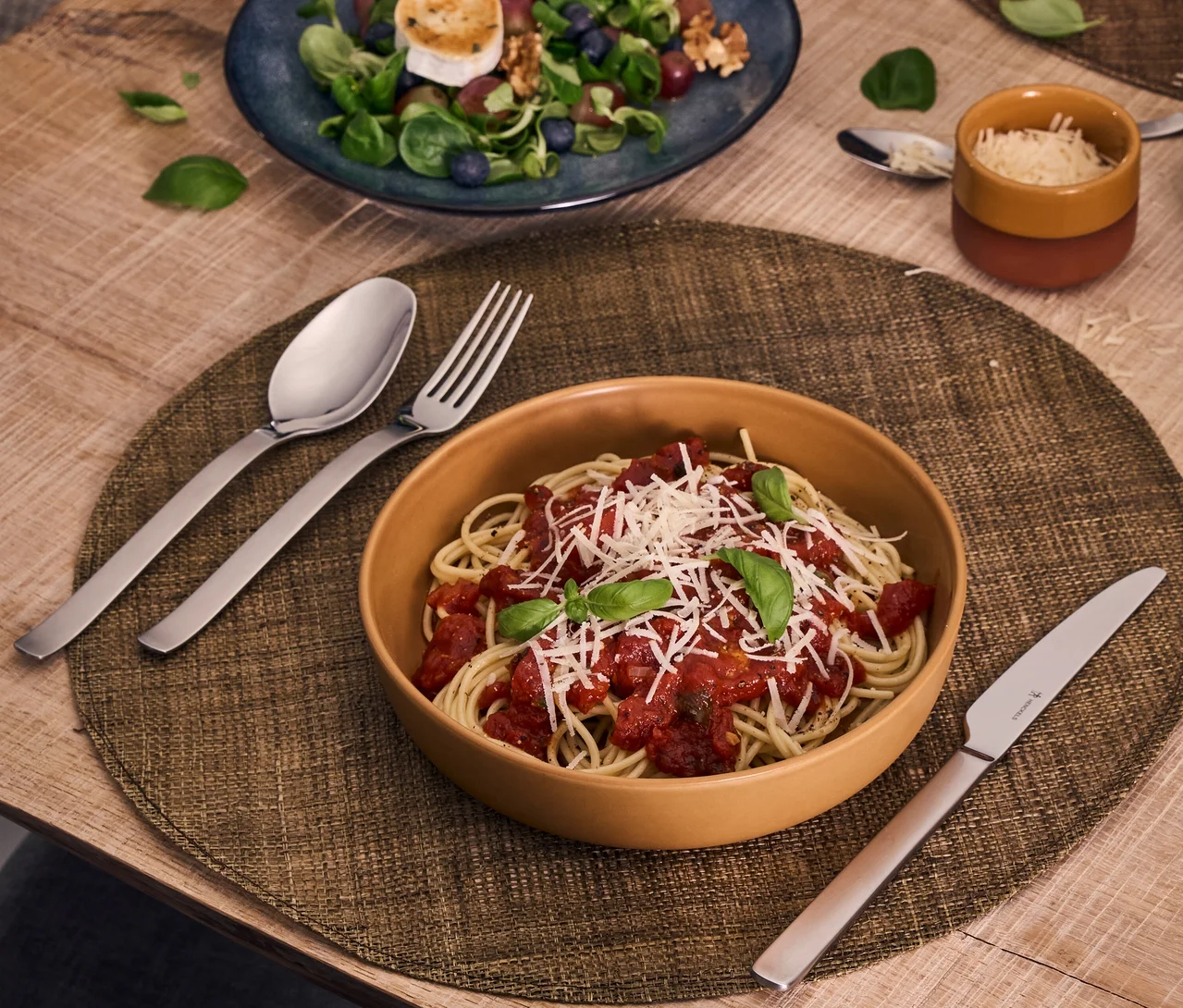 Eine Schüssel Spaghetti mit Tomatensauce, geriebenem Käse und Basilikum steht auf einem Platzset, umgeben von einem Henckels by ZWILLING Clouds Löffel, Gabel und Messer. Im Hintergrund sind ein Salat und ein Käsetopf zu sehen.
