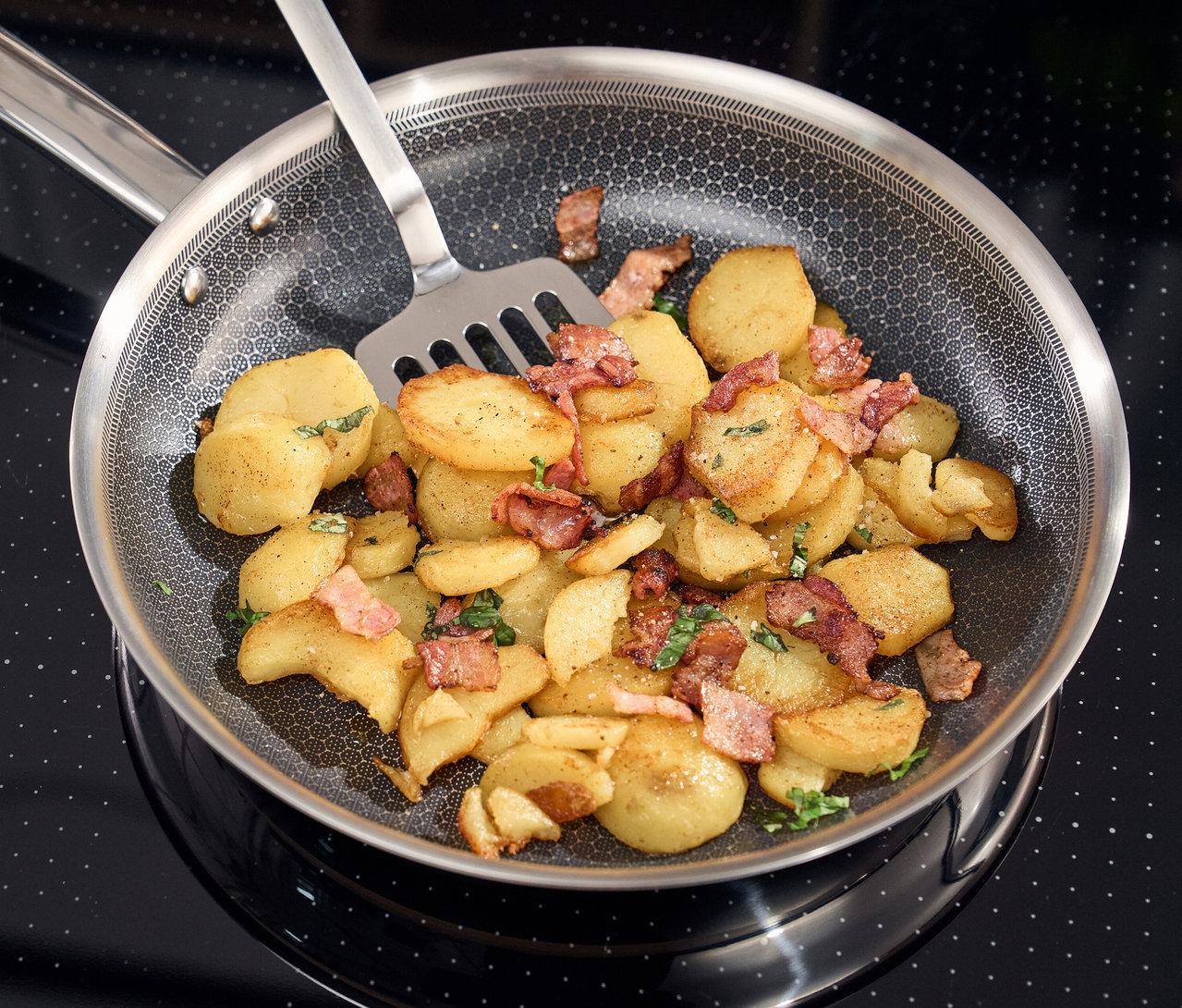 Eine Henckels by ZWILLING »Valley« Bratpfanne mit goldbraunen Kartoffeln und knusprigem Speck auf einem schwarzen Induktionskochfeld. Ein Spatel ist in der Pfanne.