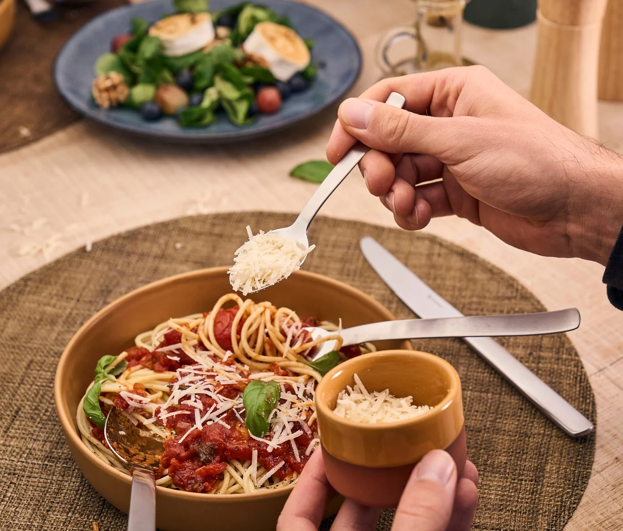 Eine Person streut geriebenen Käse von einem Löffel auf eine Schüssel Spaghetti mit Tomatensauce und Basilikum. Eine Gabel des Henckels by ZWILLING »Clouds« Bestecksets steckt in den Nudeln, ein passendes Messer liegt auf dem Tischset. Eine weitere Hand hält ein kleines Käsetöpfchen. Im Hintergrund ist unscharf ein Salat mit Ziegenkäse zu sehen.