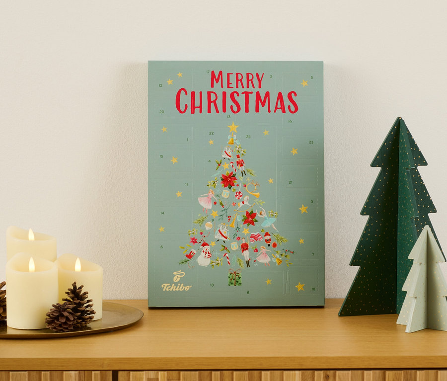 Tchibo Adventskalender auf einer Holzkommode neben Kerzen und Deko-Tannenbäumen.