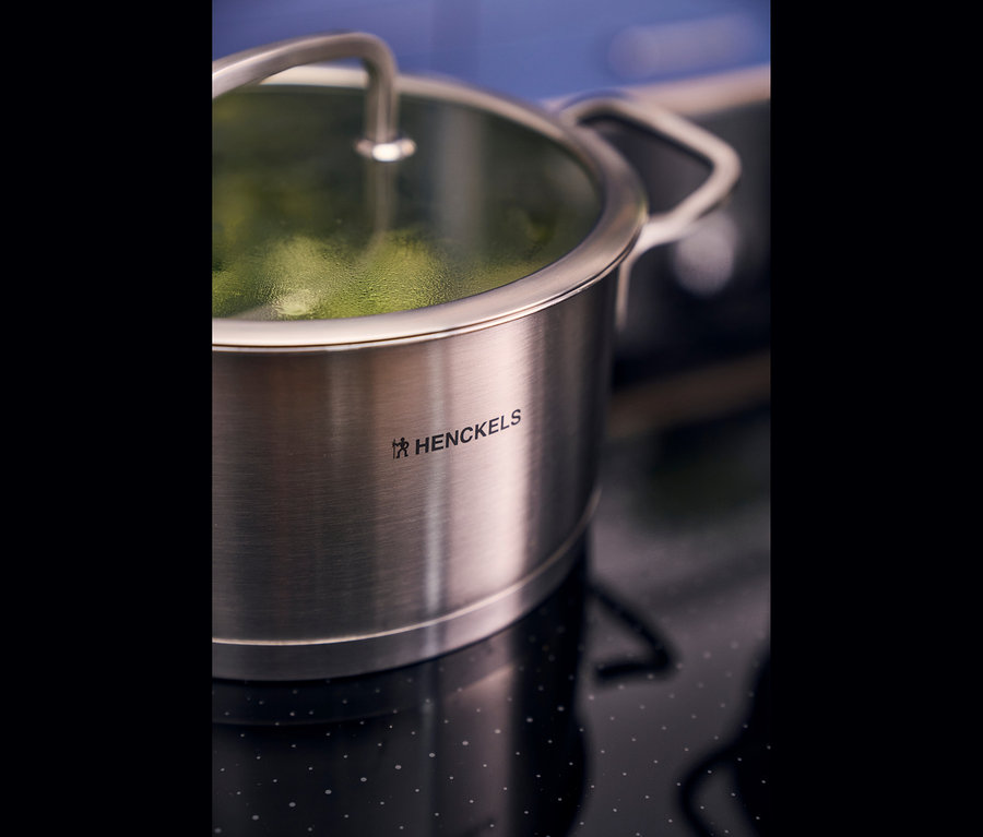 Edelstahltopf aus dem Henckels by ZWILLING »Pulse« Kochgeschirrset mit Glasdeckel auf einem schwarzen Kochfeld, in dem grünes Gemüse dämpft.