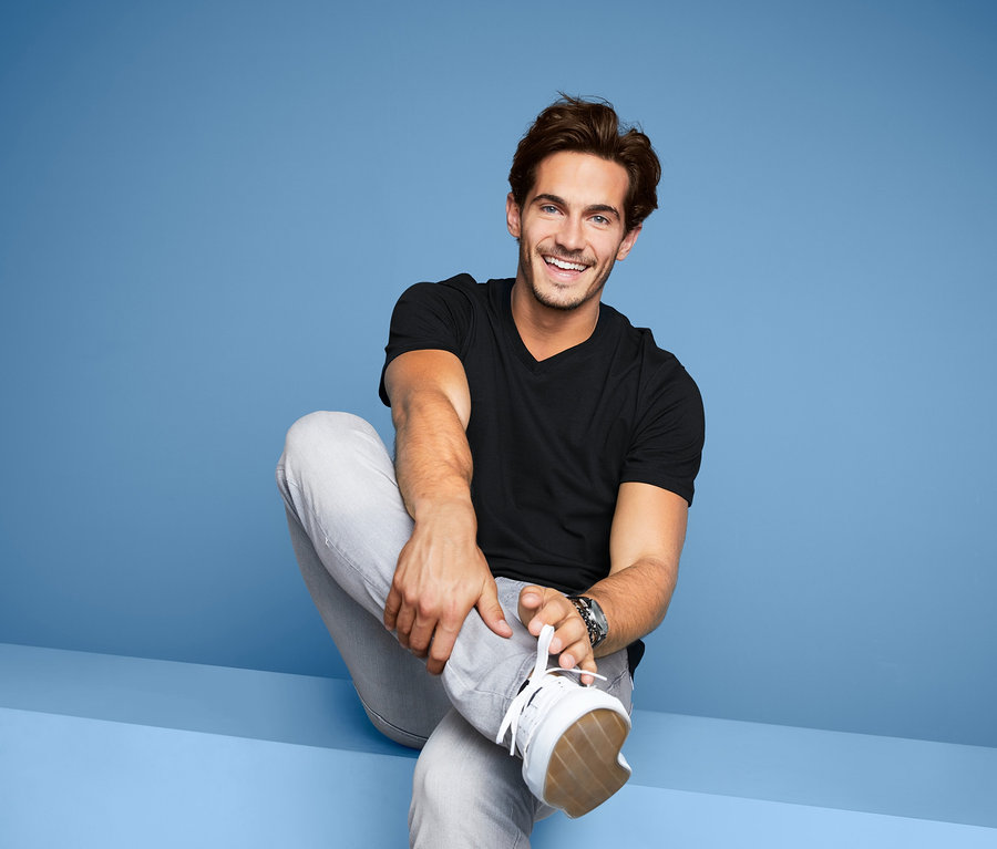 Mann sitzt lächelnd, trägt ein schwarzes 2 T-Shirts mit V-Ausschnitt, graue Jeans und Turnschuhe.