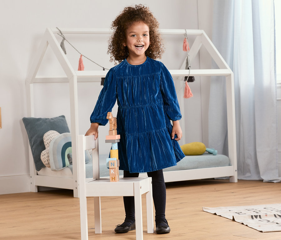 Mädchen im Samtkleid steht an einem Stuhl mit Holzbausteinen.