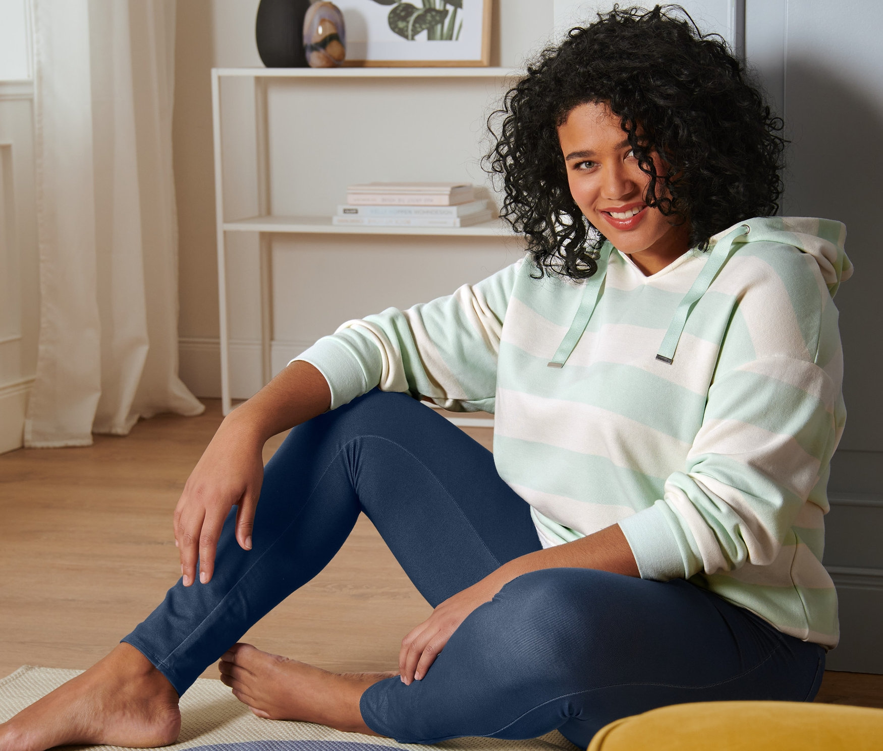 Frau sitzt auf dem Boden und trägt ein gestreiftes Kapuzen-Sweatshirt in Mint-Creme und Leggings in Jeansoptik.