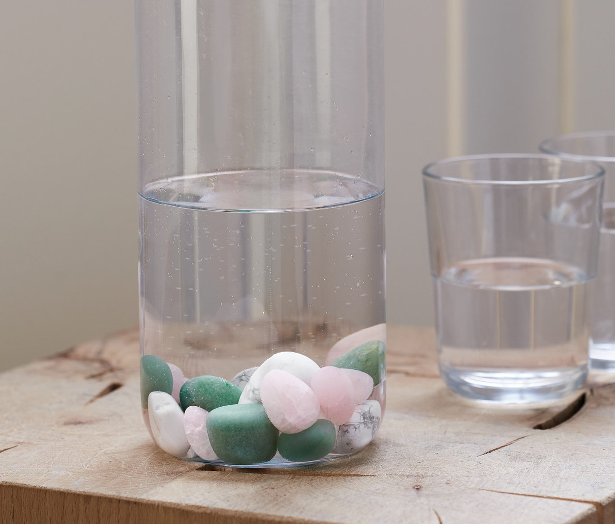 Eine klare Flasche mit Korkverschluss, teilweise gefüllt mit Wasser und Wasser-Steine, steht auf einem Holzblock neben zwei Gläsern Wasser.