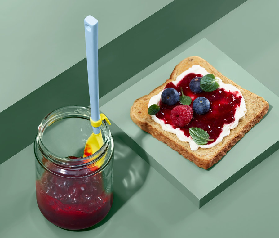 Marmelade im Glas mit blau-gelbem Spezial-Silikonlöffel und Toast mit Marmelade.