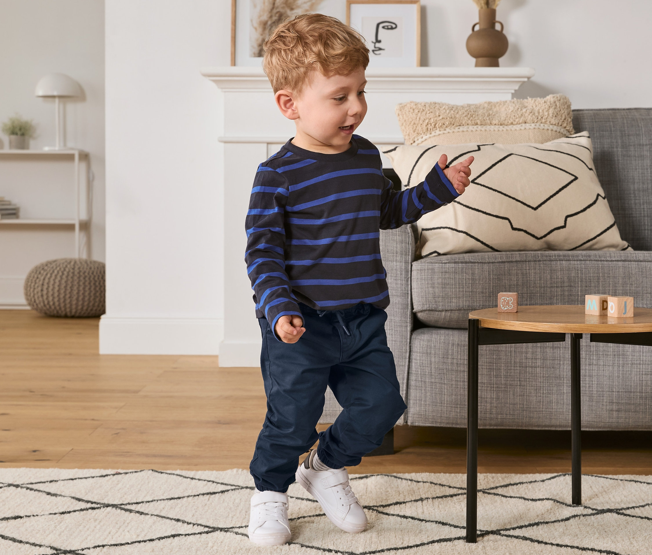 Kleiner Junge spielt mit Bauklötzen auf einem Tisch, er trägt eine blaue Chino-Schlupfhose und ein gestreiftes Hemd.