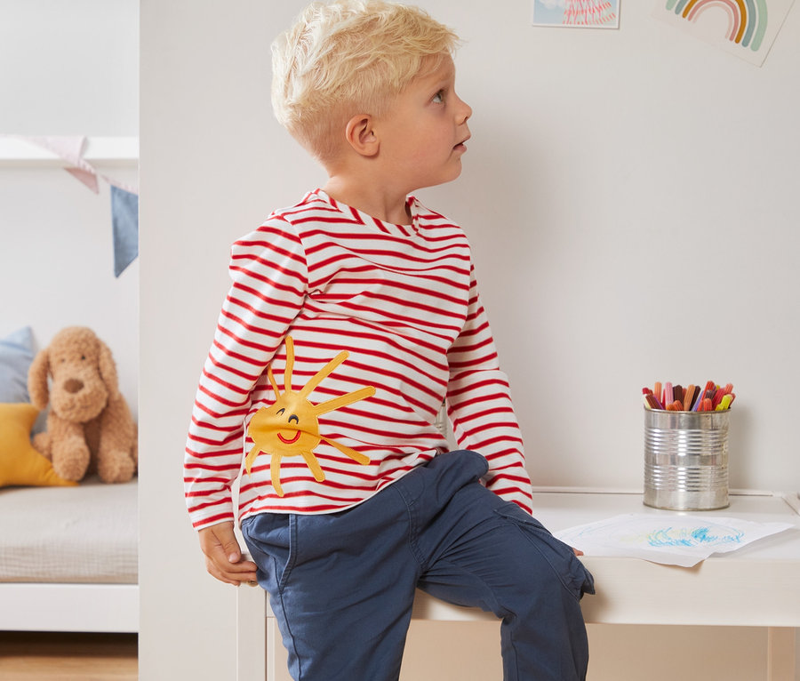 Ein Junge sitzt auf einem weißen Tisch und trägt ein rot-weiß gestreiftes Langarmshirt und blaue Hosen. Er trägt 1 von 2 Jersey-Langarmshirts aus Bio-Baumwolle.