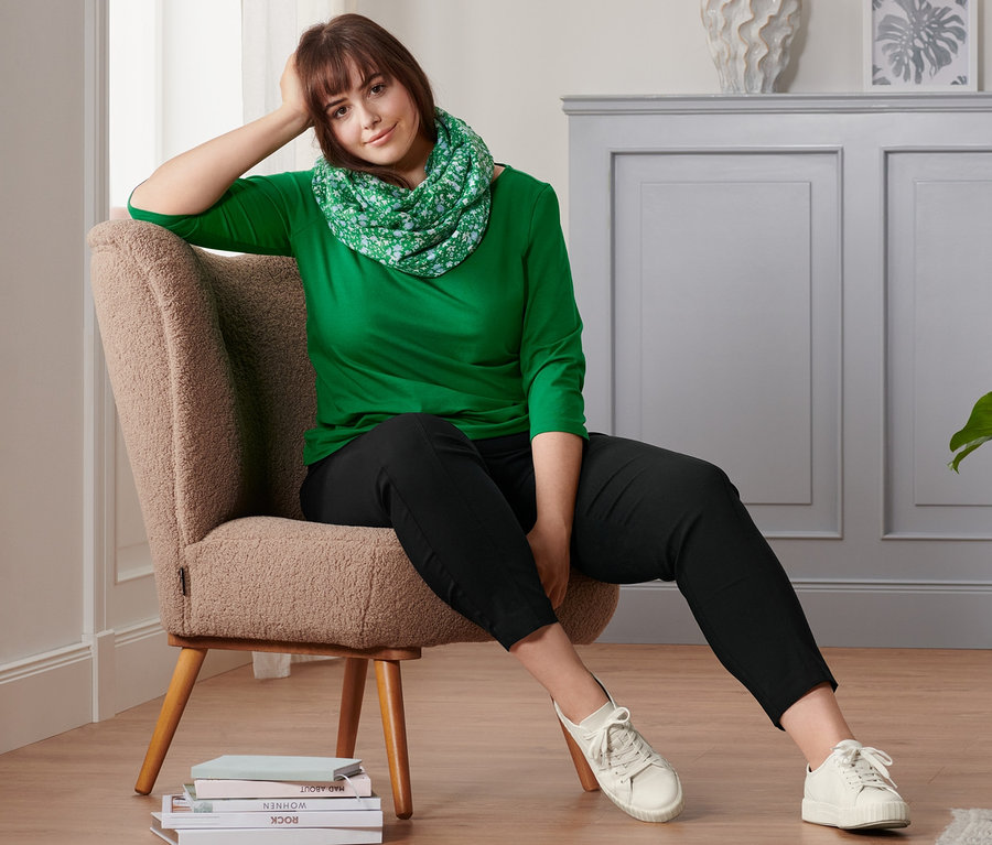 Eine Frau sitzt in einem Sessel und trägt ein grünes Shirt mit 3/4-Arm, einen Loop-Schal, eine schwarze Stretchhose und Leder-Sneaker.