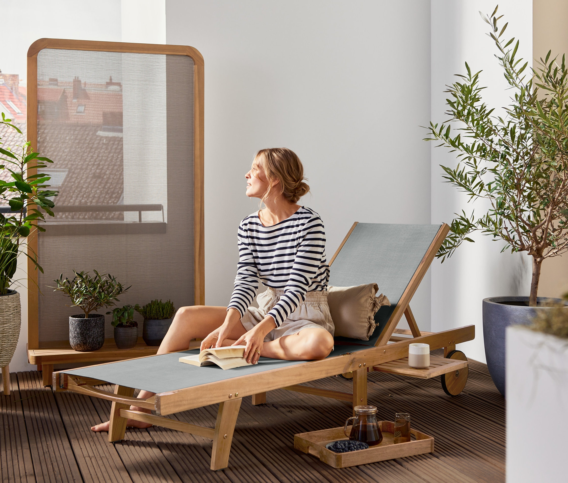 Frau sitzt auf der Liege »Lenja« mit ausziehbarem Tabletttisch und liest ein Buch. Im Hintergrund befindet sich ein Sichtschutz Textilene »Lenja«.