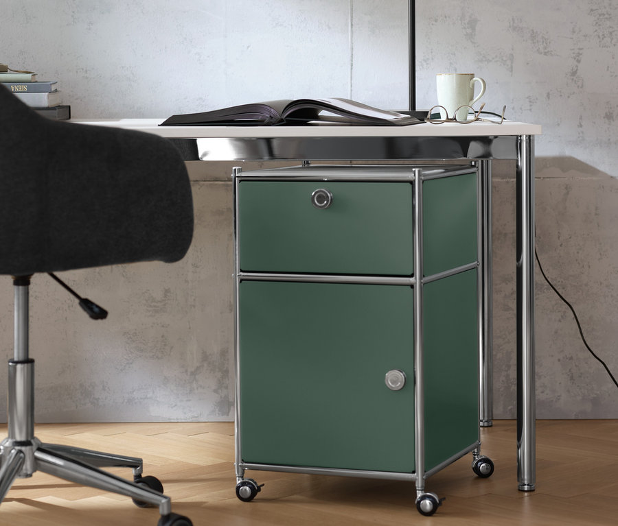 Auf einem Schreibtisch »CN3« stehen eine Tischleuchte aus Metall, ein offenes Buch, eine Brille und eine Tasse. Unter dem Tisch steht ein olivgrüner Rollcontainer »CN3« mit Schublade und Tür.