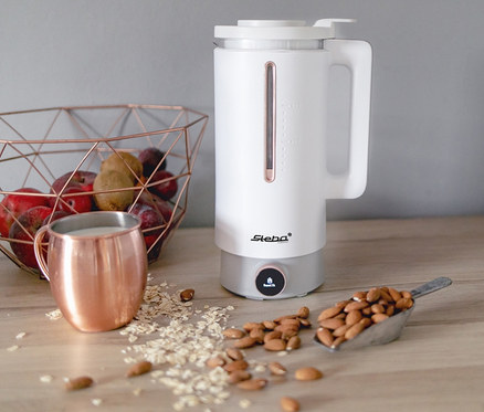 Auf einem Holztisch stehen ein weißer Steba Vegan-Drink-Maker »VDM 2 HOT & COLD«, ein Kupferbecher mit Getränk, ein Metallkorb mit Obst und ein Löffel Mandeln.