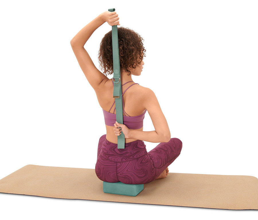 Frau sitzt auf Yogamatte und benutzt ein Yoga-Set, inklusive Block und Gurt.