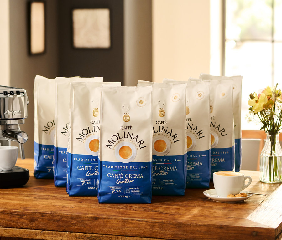 Espressomaschine mit Tasse und acht Packungen Caffè Molinari Caffè Crema Gustoso - 8x 1 kg Ganze Bohne auf einem Holztisch.