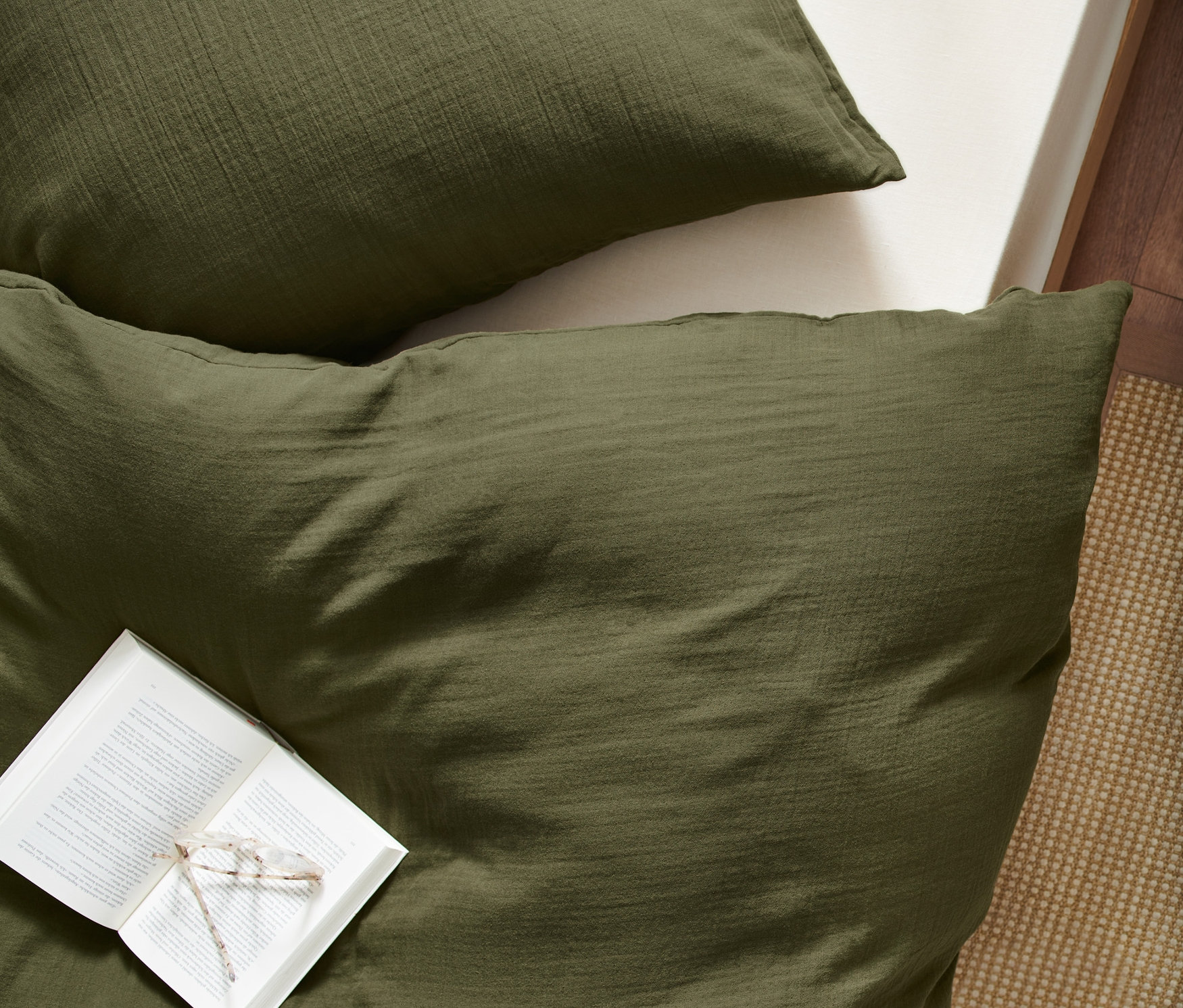 Draufsicht auf olivgrüne Musselin-Bettwäsche Normalgröße mit offenem Buch und Brille darauf.