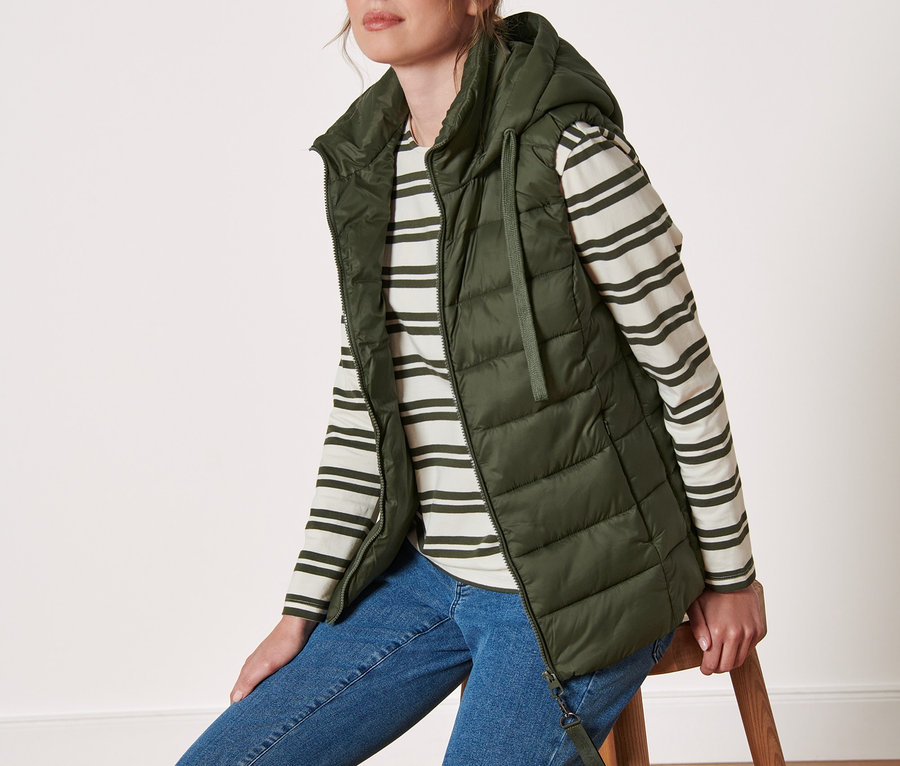 Frau sitzt auf einem Hocker und trägt eine Steppweste mit Kapuze.