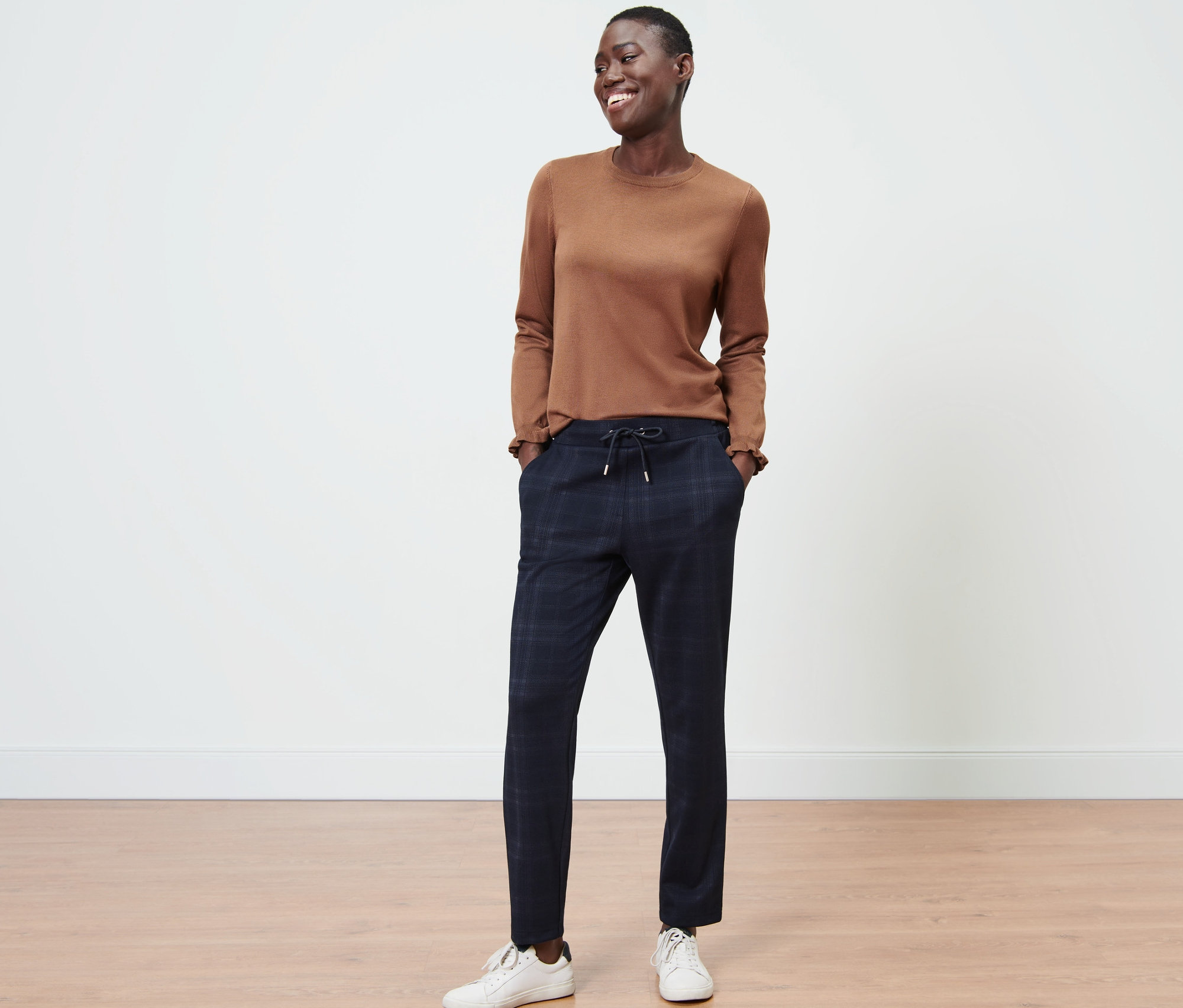Eine Frau steht und lächelt, sie trägt ein braunes Oberteil und eine karierte Joggpants.