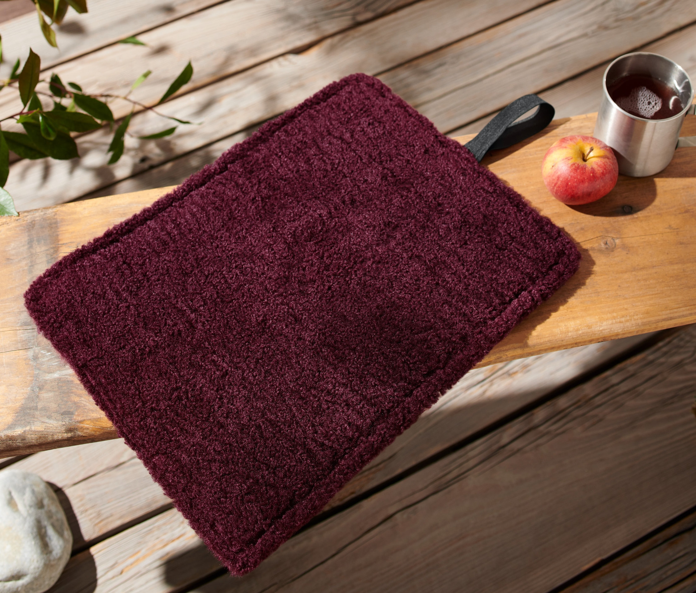 Ein faltbares Thermo-Sitzkissen in Weinrot liegt auf einer Holzbank neben einem Apfel und einer Tasse.